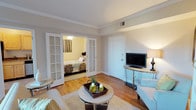 This single-floor space offers a thoughtfully designed layout that balances comfort and functionality. The living area serves as the central hub, showcasing warm hardwood flooring that extends throughout, complemented by neutral-toned walls and crisp white crown molding that adds a refined architectural detail. A plush area rug anchors the seating arrangement, centered around a round glass coffee table that contributes a modern, airy touch to the room. Built-in ceiling vents ensure efficient climate control throughout.

A standout feature of the space is the pair of classic white French doors with glass panel detailing, which elegantly connect the living area to the bedroom. The bedroom, visible through these doors, features warm lighting, a ceiling fan, and rich, patterned bedding, creating an inviting and restful retreat. The contrast between the brighter living space and the warmer tones of the bedroom adds a pleasant sense of depth and variety.

A console table along one wall provides practical surface space, and overall, the layout flows naturally from one area to the next, making the space feel cohesive, well-appointed, and comfortable for everyday living.