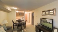 This single-floor residence offers a practical and comfortable open-concept layout that seamlessly connects the living, dining, and kitchen areas. The living area provides ample space for relaxation and entertainment, anchored by a media console and flat-screen television, with warm, neutral wall tones contributing to a cozy atmosphere throughout.

Adjacent to the living area, the dining space accommodates a full-sized dining table with seating for four, making it well-suited for both everyday meals and casual gatherings. A framed wall accent adds a decorative touch to the space.

The kitchen is positioned at the rear of the open-plan area and features wood-toned upper cabinetry, a microwave, and countertop appliances, providing a functional cooking environment. A wall-mounted clock adds a classic, homey detail to the kitchen backdrop.

Soft, neutral carpeting runs throughout the main living and dining areas, enhancing the warmth of the interior. A hallway leads to the remaining rooms of the unit, including what appears to be a bedroom with a closet featuring sliding doors. The textured ceiling and flush-mount lighting fixtures are consistent throughout, providing even, ambient illumination across the space. Overall, the layout is efficient and thoughtfully arranged for comfortable day-to-day living.