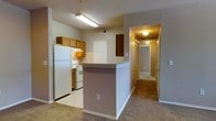 This single-floor space is centered around a functional, open-concept kitchen and living area. The kitchen is equipped with white appliances, including a refrigerator and electric range, complemented by warm wood-toned cabinetry that extends to the upper walls for generous storage. A peninsula with a neutral countertop defines the boundary between the kitchen and the adjoining carpeted area, providing both additional workspace and a natural division of the living space. Overhead, flush-mounted and fluorescent lighting keeps the area well-illuminated. A paneled white door off the kitchen leads to what appears to be a pantry or utility closet, adding practical storage. A dark wood cabinet panel provides a subtle contrast to the lighter surroundings. From the kitchen, a carpeted hallway extends toward additional rooms, lined with ceiling-mounted light fixtures that create a warm, inviting path. The neutral wall tones throughout lend a clean, understated ambiance, while the transition from tile flooring in the kitchen to carpet in the hallway and adjoining spaces adds textural variety. Overall, the layout is practical and efficiently organized.