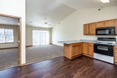 This single-floor unit is designed with an open-concept layout that seamlessly connects the kitchen and living areas. The kitchen is well-equipped with warm honey-toned oak cabinetry, a full suite of appliances including an electric range, built-in microwave, dishwasher, and refrigerator, along with a peninsula counter that provides additional workspace and separation between the kitchen and adjoining areas. The flooring in the kitchen transitions from rich dark hardwood to plush neutral carpet in the living space.

The living area benefits from vaulted ceilings that lend a sense of openness and volume, complemented by a ceiling fan overhead. A sliding glass door floods the space with natural light and provides exterior access. The overall color palette is neutral throughout, featuring cream walls and warm wood trim.

The bedrooms are carpeted and generously sized, each featuring windows for natural light. At least one bedroom includes a closet with bifold doors. The bathroom is finished with oak cabinetry, a wide vanity with a white countertop, hardwood-style flooring, and a full tub-shower combination. The unit's layout and finishes provide a clean, functional, and comfortable living environment.