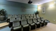 This single-floor screening room is designed to deliver a true theater experience in a private setting. The room is furnished with multiple rows of plush, reclining theater-style chairs upholstered in a patterned fabric, each equipped with built-in cup holders for added convenience. The seating is arranged in a tiered layout, ensuring unobstructed sightlines from every position in the room.

A ceiling-mounted projector serves as the centerpiece of the room's entertainment setup, positioned to project onto a dedicated screen at the front of the space. The walls are painted in a calming teal-green tone, contributing to a relaxed, cinematic ambiance. A striking metallic wall sculpture adds an artistic focal point to the back wall, while framed artwork on the side wall enhances the refined aesthetic.

Accent lighting illuminates the space warmly, complemented by a lush potted plant that introduces a natural element to the room. The light-toned flooring contrasts pleasantly with the rich upholstery of the seating. Overall, this screening room combines comfort, style, and functionality to create an inviting and immersive entertainment environment.