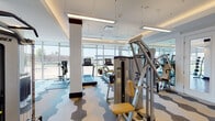 Located at 4207 E Ninth Ave in Denver, CO, this well-appointed fitness center is designed to support a comprehensive range of workout routines. The facility is organized into distinct zones across a single floor, each tailored to specific training needs. The main cardio and strength area features a bold geometric hexagonal-patterned carpet and is equipped with Cybex treadmills, ellipticals, stationary bikes, and multi-station strength machines. Floor-to-ceiling windows line the perimeter, flooding the space with natural light and offering open views of the surrounding Denver neighborhood. Recessed tray ceilings with warm accent lighting and suspended linear fixtures contribute to a clean, modern aesthetic throughout. A dedicated free weights zone transitions to smooth hardwood flooring and includes an adjustable weight bench, a full rack of dumbbells, stability balls of varying sizes, and resistance bands, accompanied by large mirrors that enhance the sense of space. An adjacent open area with hardwood floors provides room for stretching, functional training, and mat-based exercises, with additional equipment such as a stair climber accessible nearby. The facility is well-lit and thoughtfully laid out, creating an energizing and functional environment for users at all fitness levels.