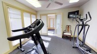 This single-floor fitness space is thoughtfully designed to provide a functional and comfortable workout environment. The room features carpet flooring and is illuminated by a combination of overhead fluorescent lighting and a ceiling fan, which helps maintain airflow during exercise sessions. Large windows dressed with blinds allow natural light to fill the space while offering privacy, contributing to a bright and welcoming atmosphere.

The fitness area is equipped with cardio equipment, including a treadmill prominently positioned in the room, making it well-suited for daily exercise routines. A wall-mounted flat-screen television is conveniently installed, allowing users to stay entertained during workouts. A traditional wooden chair with an upholstered seat is also present, providing a spot to rest or store personal items.

French-style glass-paneled doors open to a connecting hallway, enhancing the sense of openness and allowing easy access to the rest of the space. The soft yellow-toned walls lend a calm, neutral aesthetic that complements the functional purpose of the room. Overall, this fitness space strikes a balance between practicality and comfort, making it an inviting setting for regular physical activity.