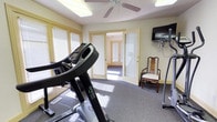 This single-floor fitness space is thoughtfully designed to provide a functional and comfortable workout environment. The room features carpet flooring and is illuminated by a combination of overhead fluorescent lighting and a ceiling fan, which helps maintain airflow during exercise sessions. Large windows dressed with blinds allow natural light to fill the space while offering privacy, contributing to a bright and welcoming atmosphere.

The fitness area is equipped with cardio equipment, including a treadmill prominently positioned in the room, making it well-suited for daily exercise routines. A wall-mounted flat-screen television is conveniently installed, allowing users to stay entertained during workouts. A traditional wooden chair with an upholstered seat is also present, providing a spot to rest or store personal items.

French-style glass-paneled doors open to a connecting hallway, enhancing the sense of openness and allowing easy access to the rest of the space. The soft yellow-toned walls lend a calm, neutral aesthetic that complements the functional purpose of the room. Overall, this fitness space strikes a balance between practicality and comfort, making it an inviting setting for regular physical activity.