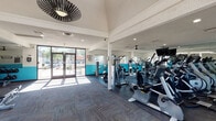 This single-level fitness center is designed to support a comprehensive workout experience in a bright, open environment. The space is anchored by large glass entry doors on two sides, flooding the interior with natural light and offering views of the surrounding outdoor area, including a pool and landscaping. A bold two-tone color scheme — white upper walls paired with vibrant teal lower walls — gives the facility a modern, energetic aesthetic, complemented by stylish mixed-tone carpet flooring and recessed lighting throughout.

The main workout area is generously equipped with a full row of treadmills, ellipticals, and stationary bikes, all facing large wall-to-ceiling mirrors that enhance the sense of space. Ceiling fans provide additional air circulation across the cardio zone. A dedicated strength training section features a variety of cable and resistance machines, along with free weight benches and storage racks mounted on the teal accent walls.

A seating area near the entrance offers a place to rest, featuring framed artwork and a water station. Posted rules confirm the facility operates 24 hours. A striking woven pendant light fixture adds a decorative touch near the entrance, reinforcing the space's blend of function and style.
