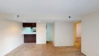This single-floor residence features two bedrooms and one and a half bathrooms within a thoughtfully arranged floor plan. The main living area is finished with neutral-toned carpet, creating a warm, cohesive atmosphere that flows naturally through the space. Recessed lighting illuminates the interior, complemented by ceiling-mounted HVAC vents that ensure year-round comfort.

The kitchen is defined by dark espresso-toned cabinetry, which provides a striking contrast against the light walls and countertops. A pass-through counter connects the kitchen to the living area, promoting an open feel while maintaining a functional separation between cooking and lounging spaces. Under-cabinet lighting adds a practical touch to the kitchen zone.

A doorway visible from the main living area leads to additional rooms, with hardwood or wood-style flooring hinting at the character found deeper in the unit. Clean white walls throughout keep the space feeling bright and versatile. The layout is efficient and straightforward, making it well-suited for everyday living with easy flow between the principal areas.