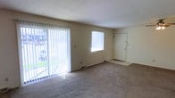 This single-floor space centers around a modest living area finished with wall-to-wall carpet in a neutral tone, lending a quiet and understated feel to the interior. The room is naturally illuminated by two sources of light: a large sliding glass door fitted with vertical blinds that opens to a view of a grassy outdoor area, and a secondary window covered by horizontal blinds on the adjacent wall. Together, these features allow ample daylight to fill the space throughout the day. The walls are finished in a light, neutral tone, complementing the simple, clean aesthetic of the room. A textured ceiling adds a subtle visual element overhead. A standard entry door is positioned along one wall, providing a clearly defined entryway with a small transition area at the threshold. A floor vent is visible near the base of the wall, indicating a forced-air climate system. The overall layout is open and unfurnished, making the space easy to visualize and configure for various uses. The design is straightforward and functional, suited to everyday living.