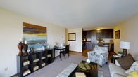 This single-floor residence offers an open-concept layout that integrates the living, dining, and kitchen areas into a unified, functional space. The living area is anchored by plush carpeting and features a stylish black shelving unit displaying decorative accessories beneath a bold, colorful abstract artwork that serves as a focal point on the neutral-toned wall. A patterned accent chair and a dark coffee table with decorative elements add character and warmth to the seating area.

Adjacent to the living space, a compact dining nook accommodates a dark bistro-style table and chairs, positioned beside a small desk area — ideal for working from home. The transition from carpet to hardwood flooring marks the entry into the kitchen, which is fitted with dark cabinetry extending to the upper walls, providing generous storage. A pass-through window connects the kitchen visually to the main living space, enhancing the open feel.

The overall ambiance is warm and inviting, with neutral walls and layered textures creating a balanced, contemporary aesthetic throughout.