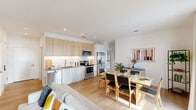 Located in Bethesda, MD, this thoughtfully designed single-floor unit offers a seamless blend of modern aesthetics and practical living. The open-plan layout connects the living, dining, and kitchen areas into one cohesive, light-filled space. Light hardwood flooring runs throughout, complementing the crisp white walls and creating a warm, airy atmosphere.

The kitchen is outfitted with stainless steel appliances — including a range, microwave, and French door refrigerator — paired with light wood upper cabinetry, white lower cabinets, a neutral-toned countertop, and a clean white tile backsplash. The dining area accommodates a full-sized table and flows naturally into the living room, which features recessed lighting and ample space for relaxed seating arrangements.

The bedroom is a calm retreat, highlighted by floor-to-ceiling windows that frame lush outdoor greenery and fill the room with natural light. Soft carpeting and neutral tones contribute to a serene ambiance. The bathroom is finished with a glass-enclosed walk-in shower featuring white tile surround and clean chrome fixtures. Throughout the unit, the design maintains a consistent contemporary sensibility — minimal, functional, and welcoming.