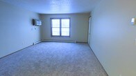 This single-floor residential unit offers a straightforward and functional layout designed for comfortable everyday living. The main living area is finished with wall-to-wall carpet in a neutral tone, providing a warm and practical underfoot surface throughout the space. A centrally placed window allows natural light to filter into the room, lending the interior a bright and open feel. Beneath the window, a baseboard heating unit runs the length of the wall, ensuring efficient climate control during colder months. Complementing the heating system, a wall-mounted air conditioning unit is positioned on an adjacent wall, offering year-round temperature management. The ceiling features a classic textured finish, adding subtle visual depth to the room. The walls are painted in a soft, neutral tone that creates a clean and versatile backdrop, suitable for a variety of interior styles. A standard interior door provides access to additional areas of the unit. Overall, the space presents a practical and well-equipped interior that is move-in ready, with essential climate control features and a clean, unfussy design throughout.