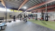 This expansive single-floor exercise facility is housed within an industrial-style structure featuring a corrugated metal roof supported by bold steel I-beams, lending the space a rugged, warehouse aesthetic. The generous floor area is thoughtfully divided into multiple functional training zones, providing ample room for a wide range of workout disciplines.

The strength training area features polished concrete flooring and is well-equipped with power racks, barbells, weight plates, adjustable benches, and an extensive dumbbell rack. A separate weightlifting platform zone, enclosed by wire mesh partitions, offers dedicated spaces for Olympic lifting with proper flooring pads and loaded barbells. Natural light filters in through openings along the upper walls, complementing the overhead industrial lighting throughout.

A large turf-covered functional fitness zone anchors the center of the facility, complete with suspended heavy bags for combat training, battle ropes, medicine ball racks, kettlebells, and a variety of conditioning tools including tires and sleds. The open layout of this area supports group training and high-intensity workouts.

Wire mesh cage dividers thoughtfully segment the space without sacrificing the open, airy atmosphere created by the soaring ceiling height. The overall environment is utilitarian, purpose-built, and well-organized, making it a fully equipped destination for serious athletic training.