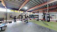 This expansive single-floor exercise facility is housed within an industrial-style structure featuring a corrugated metal roof supported by bold steel I-beams, lending the space a rugged, warehouse aesthetic. The generous floor area is thoughtfully divided into multiple functional training zones, providing ample room for a wide range of workout disciplines.

The strength training area features polished concrete flooring and is well-equipped with power racks, barbells, weight plates, adjustable benches, and an extensive dumbbell rack. A separate weightlifting platform zone, enclosed by wire mesh partitions, offers dedicated spaces for Olympic lifting with proper flooring pads and loaded barbells. Natural light filters in through openings along the upper walls, complementing the overhead industrial lighting throughout.

A large turf-covered functional fitness zone anchors the center of the facility, complete with suspended heavy bags for combat training, battle ropes, medicine ball racks, kettlebells, and a variety of conditioning tools including tires and sleds. The open layout of this area supports group training and high-intensity workouts.

Wire mesh cage dividers thoughtfully segment the space without sacrificing the open, airy atmosphere created by the soaring ceiling height. The overall environment is utilitarian, purpose-built, and well-organized, making it a fully equipped destination for serious athletic training.