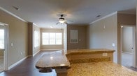 Located at 1001 Quarters in Sneads Ferry, NC, this single-story home offers a comfortable and well-appointed living experience. The open-concept layout creates a seamless flow between the kitchen and living areas, making the space feel expansive and welcoming. The kitchen is highlighted by striking granite countertops with warm golden and amber tones, providing both elegance and durability. A raised breakfast bar adds a functional yet stylish element, ideal for casual dining or entertaining. The living area features classic crown molding that frames the ceilings and adds a refined architectural touch throughout. A ceiling fan with a light fixture keeps the space comfortable year-round while complementing the room's traditional aesthetic. Large windows along one wall invite generous amounts of natural light, creating a bright and airy atmosphere. Hardwood-style flooring runs through the common areas, adding warmth underfoot and tying the interior together cohesively. Neutral wall tones provide a versatile backdrop that complements a variety of décor styles. The home's single-floor layout ensures easy accessibility and practical living, making it a functional and inviting residence in the Sneads Ferry community.