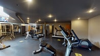 Located at 505 N Peoria St in Chicago's West Town neighborhood, this fitness center is designed to accommodate a comprehensive range of workout needs. The space features a striking industrial aesthetic, highlighted by soaring double-height ceilings with an exposed upper mezzanine level bordered by metal railings. Bold yellow accent walls contrast with dark, neutral tones throughout, creating an energetic yet focused atmosphere. Recessed and pendant lighting illuminate the rubber-floored workout areas effectively.

The main floor is organized into distinct training zones. A dedicated cardio section houses multiple treadmills and a stationary bike, while a strength training area features a power rack, Smith machine, and multi-station cable machine suitable for a wide variety of resistance exercises. The free weights section includes a full dumbbell rack alongside barbells, weight plates, and adjustable benches, providing ample options for both compound and isolation movements. A mounted flat-screen television displaying sports programming adds an engaging element to workouts.

Entry to the fitness floor is through a sleek, modern corridor framed by dark architectural columns and large glass panels, offering a clean and professional transition into the workout space. Upper-level windows allow natural light to filter in, complementing the overhead artificial lighting and enhancing the overall openness of the facility.