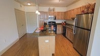 This single-floor space showcases a thoughtfully designed kitchen that serves as the heart of the home. The kitchen is anchored by a central island topped with rich dark granite countertops, offering both additional prep space and casual seating. Matching granite surfaces extend to the surrounding countertops, providing a cohesive and upscale aesthetic throughout. Warm hardwood flooring runs throughout the space, adding a natural and inviting tone to the overall environment. The cabinetry is finished in a classic honey-toned wood, featuring raised panel detailing and ample storage that extends all the way to the upper reaches of the walls, maximizing vertical space. Stainless steel appliances, including a refrigerator, microwave, and dishwasher, complement the darker black range, creating a balanced contrast against the lighter cabinetry. Overhead, a combination of pendant lighting above the island and flush-mount ceiling fixtures provides layered illumination suited to both task and ambient needs. Track lighting further enhances the functionality of the cooking area. The space feels open and well-proportioned, with a smooth flow between the cooking and prep zones, making it both practical and visually appealing for everyday use.