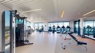 This contemporary fitness center is designed to support a comprehensive range of workout disciplines across multiple interconnected zones. The main cardio and strength area is expansive and open, featuring durable rubberized flooring and an array of equipment including treadmills, elliptical trainers, cable machines, weight benches, and a full dumbbell rack. Floor-to-ceiling windows line the perimeter, flooding the space with abundant natural light and offering sweeping panoramic views of the surrounding skyline and landscape, creating an energizing atmosphere for training.

A separate studio space adjoins the main floor, featuring smooth white flooring, full-length mirrored walls, and a mounted flat-screen display — ideal for group classes, stretching, or personal training sessions. Step platforms and free weights are available within this room, which also benefits from direct access to natural light through large glass doors.

An additional cycling zone provides a quieter, focused environment for indoor cycling workouts, equipped with stationary bikes and a monitor, with continued access to the expansive views through the surrounding glass walls.

Throughout the facility, recessed and linear LED lighting complements the natural light, while the crisp white walls and modern architectural finishes contribute to a clean, professional aesthetic. The layout is thoughtfully organized to allow for simultaneous use of multiple activity areas without congestion.