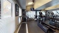 This single-level fitness center is thoughtfully designed to support a comprehensive workout experience. The space is outfitted with a diverse range of equipment, including a multi-function cable/weight machine, a treadmill, an elliptical trainer, a recumbent bike, and an assortment of medicine balls displayed along a mirrored wall — providing options for both cardio and strength training.

The room features durable, dark carpeted flooring suited for active use, while the walls are painted in a cool gray tone that gives the space a clean, modern aesthetic. Large windows fitted with horizontal blinds line multiple walls, allowing ample natural light to filter in while offering views of surrounding greenery. A ceiling fan and ventilation fixtures ensure proper airflow throughout.

A wall-mounted flat-screen TV is positioned for visibility across the exercise floor, adding an entertainment element to workouts. Full-length mirrors along one wall enhance the sense of space and allow users to monitor their form during exercise. A set of elegant French doors with glass panes connects the fitness room to an adjacent corridor, contributing a refined architectural detail to the otherwise functional space. Motivational artwork displayed on the walls adds personality and energy to the overall environment.