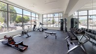 This single-floor fitness center is designed with both functionality and openness in mind. The layout is generously proportioned, offering ample floor space for a diverse range of workout activities without feeling crowded. A defining architectural feature is the expansive floor-to-ceiling window wall that spans a significant portion of the space, flooding the interior with abundant natural light and providing unobstructed views of the surrounding streetscape and neighborhood.

The flooring consists of durable rubber or carpet-style athletic material throughout, appropriate for both cardio and strength training use. Along one wall, large mirrors extend the visual depth of the room and support form-checking during exercise. The ceilings are elevated, contributing to an airy, open ambiance.

Equipment is thoughtfully arranged across the space. The cardio zone, positioned near the windows, includes treadmills, elliptical trainers, stationary bikes, and rowing machines. The strength area features a multi-station cable machine, an adjustable weight bench, and a well-stocked dumbbell rack. Additional functional training accessories, including medicine balls and resistance tools, are stored along the walls. The clean white walls and modern finishes give the space a contemporary, professional atmosphere.