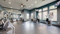 This fitness center offers a comprehensive workout environment designed to accommodate a wide range of exercise needs. The space is finished with durable rubber flooring throughout, providing a safe and functional surface suited for both cardio and strength training activities.

The main workout area is divided into distinct zones. The cardio section features multiple treadmills and an elliptical machine, positioned along a wall of large windows that invite generous natural light and offer views of surrounding greenery. A ceiling fan overhead ensures comfortable airflow during workouts.

The strength training area is equally well-appointed, housing a multi-station cable machine, a rowing machine, a stationary bike, adjustable weight benches, and a dumbbell rack. An expansive floor-to-ceiling mirror runs along one wall, visually expanding the space and allowing users to monitor their form during exercise.

Wall-mounted flat-screen televisions are positioned throughout the fitness center, offering entertainment from multiple vantage points. Recessed lighting supplements the natural light, ensuring the space remains bright and well-lit at all hours.

The interior design features cool blue-gray walls accented with dark wood crown molding, lending the space a polished and inviting aesthetic. The overall atmosphere is clean, organized, and purpose-built for an effective fitness experience.