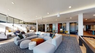 This contemporary amenity floor is thoughtfully designed to accommodate a wide range of activities, seamlessly blending comfort, productivity, and entertainment across one expansive level. Soaring double-height ceilings and floor-to-ceiling windows flood the space with abundant natural light, offering open views of the surrounding exterior and creating an airy, welcoming atmosphere throughout.

The main lounge area anchors the space with generous modular sectional sofas, eclectic ottomans, and patterned area rugs over hardwood flooring, providing a comfortable setting for casual gatherings or relaxed downtime. Adjacent co-working zones feature individual seating, task lighting, and a more focused atmosphere suited for productivity.

Glass-walled conference and private rooms line one side of the floor, framed by dark metal accents and textured wall coverings, offering transparent yet enclosed spaces ideal for meetings or quiet work sessions. A well-appointed kitchenette showcases teal cabinetry, a white tile backsplash, butcher block countertops, and a floating wood shelf displaying curated artwork.

The game room stands out with its energetic "Play. Chill. Focus." wall mural, a pool table, classic pinball machine, arcade games, and a large-screen TV — all set against the backdrop of expansive glazing. Throughout, the design strikes a balance between modern aesthetics and functional versatility.