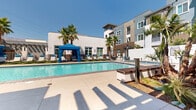 Located at 13250 Foothill Blvd in Rancho Cucamonga, California, this outdoor amenity space offers a resort-inspired environment designed for relaxation and social gatherings under the Southern California sun. At the heart of the space is a generously sized swimming pool with crystal-clear water, complemented by an adjacent spa, both finished with classic blue tile accents and wide-entry steps for easy access. Surrounding the pool, a spacious concrete deck provides ample room for lounging, lined with chaise lounge chairs and multiple blue canopied cabanas with draped curtains that offer shaded retreats. Tall, mature palm trees are strategically placed throughout, enhancing the tropical, resort-like atmosphere. A pergola-covered outdoor lounge area features comfortable seating arrangements, creating a welcoming space for socializing. An outdoor kitchen equipped with built-in grills serves as a dedicated space for al fresco dining and entertaining. The surrounding multi-story residential building, finished in a contemporary palette of white stucco and warm earth tones, frames the courtyard and adds an architectural backdrop to the amenity area. The overall design balances modern aesthetics with functional outdoor living, making it an inviting communal destination year-round.