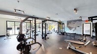 Located at 1113 Powers Ferry Pl SE in Marietta, Georgia, this fitness center offers a generously sized, single-floor workout facility designed to accommodate a comprehensive range of exercise routines. The space is thoughtfully laid out to maximize functionality, housing an extensive selection of TRUE-brand strength machines, cable systems, and squat racks alongside a full dumbbell rack and weight benches for free weight training. The cardio zone features multiple treadmills and elliptical machines positioned near floor-to-ceiling windows that look out onto an outdoor pool area, flooding the space with natural light and providing an energizing view during workouts. The durable rubber flooring spans the entire facility, providing safety and comfort underfoot, while recessed track lighting ensures consistent, bright illumination throughout. A striking, large-scale abstract mural in muted tones of yellow, gray, and blue anchors one wall, lending a modern and vibrant aesthetic to the environment. Full-length mirrors along one section of the wall enhance the sense of openness and allow users to monitor their form. The overall ambiance is clean, modern, and well-maintained, reflecting a professional fitness environment suited to both casual users and dedicated athletes.