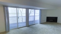 This single-floor space makes a striking first impression with its wall of floor-to-ceiling sliding glass doors that span the main living area, inviting an abundance of natural light and framing sweeping views of a tranquil waterfront and surrounding mature trees. The generous living area is finished with wall-to-wall carpeting, offering a comfortable and cohesive feel underfoot. The sliding doors provide seamless access to a private outdoor balcony, complete with a classic white railing, making it an ideal spot to take in the serene, natural scenery beyond. The open layout of the main living space is enhanced by clean, neutral-toned walls and a straightforward design that prioritizes the views as the central feature of the room. The integration of the indoor and outdoor spaces through the expansive glazing creates an airy, light-filled ambiance throughout. Whether admiring the wooded waterfront landscape or enjoying the flow of natural light across the living area, this space offers a peaceful and scenic environment well-suited for relaxed, everyday living.