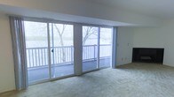 This single-floor space makes a striking first impression with its wall of floor-to-ceiling sliding glass doors that span the main living area, inviting an abundance of natural light and framing sweeping views of a tranquil waterfront and surrounding mature trees. The generous living area is finished with wall-to-wall carpeting, offering a comfortable and cohesive feel underfoot. The sliding doors provide seamless access to a private outdoor balcony, complete with a classic white railing, making it an ideal spot to take in the serene, natural scenery beyond. The open layout of the main living space is enhanced by clean, neutral-toned walls and a straightforward design that prioritizes the views as the central feature of the room. The integration of the indoor and outdoor spaces through the expansive glazing creates an airy, light-filled ambiance throughout. Whether admiring the wooded waterfront landscape or enjoying the flow of natural light across the living area, this space offers a peaceful and scenic environment well-suited for relaxed, everyday living.