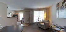Located at 6785 Oakton Ln in Columbus, Ohio, this two-floor residential space offers a welcoming and functional living environment characterized by soft neutral tones and a comfortable, understated aesthetic. The living area is a highlight of the home, featuring large windows dressed with white blinds and sheer curtains that allow generous amounts of natural light to fill the room. The space is finished with plush wall-to-wall carpeting in a warm beige tone, contributing to an inviting atmosphere. A cozy armchair and accent side table with a small floral arrangement add a homey touch, while a floor lamp provides warm supplemental lighting. The entryway is clearly defined, with a paneled front door accompanied by a small entry mat, creating a natural transition into the main living space. An additional window near the entry further brightens the area. The layout feels open and airy, with thoughtful furniture placement that maximizes usability without overcrowding the room. Subtle architectural details, including a textured ceiling and clean wall lines, lend a classic, timeless character to the interior. Spanning two floors, this space offers room for a variety of living arrangements, making it a practical and comfortable home in a Columbus neighborhood.