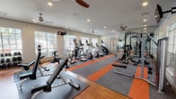 This fitness center is a spacious, single-floor facility designed to support a comprehensive workout experience. The layout is open and well-organized, with distinct zones dedicated to cardio, strength training, and free weights. The flooring is a striking combination of hardwood, gray carpet, and bold orange accent strips, giving the space a modern and energetic aesthetic.

The cardio zone is lined along the large, multi-pane windows that flood the room with natural light and offer views of the surrounding greenery. Equipment includes multiple treadmills, ellipticals, stair climbers, and stationary bikes, many outfitted with individual digital display screens. Ceiling fans positioned throughout the facility ensure consistent air circulation and comfort during workouts.

The strength training area features a power rack with weight plates and a flat bench, complemented by a multi-use cable machine. Full-length mirrors run along one wall, enhancing the sense of space and allowing users to monitor their form. The free weight section includes a dumbbell rack with an adjustable bench nearby.

Recessed lighting combined with the abundance of natural light from the large windows creates a bright, motivating atmosphere. Glass-paned doors provide access to the outdoors, further connecting the interior to its surroundings. Overall, the fitness center is thoughtfully appointed with a broad range of equipment to accommodate various fitness goals and training styles.