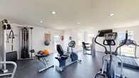 This fitness center is designed to support a well-rounded workout experience within a clean, single-floor layout. The space is finished with crisp white walls and durable dark rubber flooring, creating a practical yet inviting environment. Recessed lighting throughout ensures consistent, even illumination, while multiple windows on several walls invite ample natural light and provide views of the surrounding exterior.

The main workout area houses a comprehensive selection of cardio equipment, including treadmills, upright and recumbent stationary bikes, and an elliptical trainer — all from the True brand. A dedicated strength-training section features a multi-function cable machine, a weight bench, and a full rack of dumbbells in varying sizes. Medicine balls and stability balls are neatly stored along the walls, offering additional functional training options. A wall-mounted medicine ball rack keeps these accessories organized and accessible.

A split-unit air conditioning system ensures year-round climate comfort, while a wall-mounted flat-screen television adds an entertainment option for cardio sessions. Safety features include a visible fire extinguisher near the entry door, which offers direct outdoor access with natural light streaming through its glass panel.