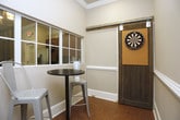 This well-appointed single-floor space combines functional workspace with leisure amenities, creating a versatile environment suited for both productivity and entertainment. Throughout the space, warm cork flooring provides a distinctive and comfortable underfoot experience, complemented by crisp white wainscoting, crown molding, and recessed lighting that lend a polished, refined character to every room.

The office area is a standout feature, thoughtfully designed with built-in shelving and an open, airy layout. Frosted glass partition panels frame the workspace, allowing natural light to filter through while maintaining a sense of privacy. The room is spacious enough to accommodate multiple workstations, making it well-suited for collaborative or individual work.

Adjacent to the office, a dedicated game room offers a fun and relaxed retreat. The room is highlighted by a rustic sliding barn door mounted with a dartboard, adding a charming, industrial-farmhouse touch to the space. A high-top bistro table with metal stools provides casual seating, and an interior window connects the game room visually to the surrounding areas, enhancing the open, communal feel of the overall layout.

Together, these spaces deliver a cohesive blend of practicality and recreation, making this an engaging and functional environment.
