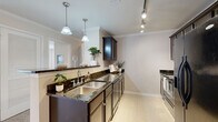 This single-floor, 2-bedroom, 2-bathroom residence offers a thoughtfully designed layout with a blend of functional and aesthetic elements throughout. The galley-style kitchen is a standout feature, showcasing rich dark espresso cabinetry, dark granite countertops with striking veining, a stainless steel double-basin sink, and stainless steel appliances including a full-size refrigerator and range. Pendant lighting and a track lighting system illuminate the space, while light tile flooring and crown molding add a refined touch. The open pass-through connects the kitchen to the dining area, enhancing the sense of flow. The living room is carpeted and features crown molding, a chandelier, and a tray ceiling detail that adds architectural interest. The spacious primary bedroom includes plush carpeting and a large window with blinds that invites natural light and outdoor views of surrounding greenery. The second bedroom is similarly carpeted and bright with neutral tones. Both bathrooms are finished with tile flooring, white vanity cabinetry, countertop sinks, and a shower-tub combination. A dedicated closet provides additional storage. The overall palette is neutral and warm, creating a clean and inviting atmosphere throughout.