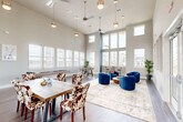 Located at 515 Dunlop Blvd SW in Madison, Alabama, this single-floor community amenity space offers a generously sized, open-plan layout designed for socializing and recreation. The room immediately impresses with its dramatic double-height ceilings, which are adorned with multiple pendant lights and ceiling fans that complement the bright, airy atmosphere created by an expansive array of large windows on multiple walls. Natural light floods every corner of the space, offering views of the surrounding community.

The interior is thoughtfully divided into distinct functional zones. A lounge area features bold blue velvet accent chairs arranged around a central rug, providing a stylish setting for casual gatherings. Nearby, a rustic wood dining table paired with cowhide-patterned chairs offers a welcoming spot for shared meals or group activities. A dedicated games area includes both a foosball table and a shuffleboard table, catering to recreational interests. A quieter seating nook, furnished with leather rocking chairs and a wall-mounted display, provides a relaxed retreat within the space.

Dark hardwood-style flooring runs throughout, grounding the neutral gray wall palette accented by a striking teal blue column. Double glass entry doors with sidelights add to the open, welcoming feel of this well-appointed communal space.
