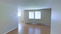 This single-floor space offers a clean, functional layout with an open and airy feel. The living area is finished with warm hardwood flooring that reflects natural light, contributing to a bright and welcoming atmosphere. Neutral-toned walls provide a versatile backdrop suitable for a variety of furnishing styles.

Large windows fitted with horizontal blinds allow for ample natural light while offering privacy control. The windows are positioned to maximize daylight throughout the main living area, creating a well-lit environment at all hours of the day. Baseboard heating units run along the walls, ensuring efficient and consistent warmth throughout the space.

The layout flows openly between areas, with a clear sightline from the main living space into an adjoining room, suggesting a practical and connected floor plan. White trim and baseboards complement the neutral wall tones, adding a crisp, finished look to the interior. The overall design is straightforward and understated, prioritizing comfort and usability. The space presents a solid foundation for personalization, making it well-suited for a range of occupants or uses.