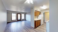 This single-floor space offers a practical and comfortable open-concept layout that seamlessly connects the living area and kitchen. The living area is finished with smooth hardwood-style flooring and features a ceiling fan with integrated lighting, providing both comfort and function. Two large windows alongside a set of glass balcony doors allow natural light to fill the room while offering direct access to an outdoor balcony, bringing a sense of openness to the interior. The kitchen is defined by warm-toned wood cabinetry — both upper and lower — paired with light countertops that provide ample workspace. A full-sized refrigerator anchors the kitchen, and the layout is efficient and well-organized. A textured ceiling adds subtle visual character throughout the main living space. The flooring carries consistently through both the living and kitchen areas, reinforcing the cohesive, open feel of the floor plan. A hallway visible from the kitchen leads to additional areas of the unit, suggesting a well-structured layout with dedicated private spaces. The overall ambiance is clean, functional, and move-in ready, making it a solid and straightforward living space.
