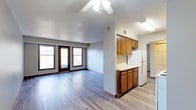 This single-floor space offers a practical and comfortable open-concept layout that seamlessly connects the living area and kitchen. The living area is finished with smooth hardwood-style flooring and features a ceiling fan with integrated lighting, providing both comfort and function. Two large windows alongside a set of glass balcony doors allow natural light to fill the room while offering direct access to an outdoor balcony, bringing a sense of openness to the interior. The kitchen is defined by warm-toned wood cabinetry — both upper and lower — paired with light countertops that provide ample workspace. A full-sized refrigerator anchors the kitchen, and the layout is efficient and well-organized. A textured ceiling adds subtle visual character throughout the main living space. The flooring carries consistently through both the living and kitchen areas, reinforcing the cohesive, open feel of the floor plan. A hallway visible from the kitchen leads to additional areas of the unit, suggesting a well-structured layout with dedicated private spaces. The overall ambiance is clean, functional, and move-in ready, making it a solid and straightforward living space.