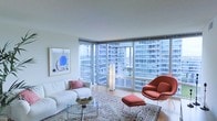 This contemporary single-floor residence offers a refined living experience defined by clean lines, an abundance of natural light, and stunning city views. The living area is the centerpiece of the space, anchored by dramatic floor-to-ceiling corner windows that wrap around two walls, framing a sweeping urban skyline panorama and flooding the interior with daylight throughout the day. The open layout is enhanced by smooth, light-toned flooring that complements the neutral palette of the walls and ceiling, creating a calm and airy atmosphere. The living room is thoughtfully appointed, featuring a plush white sofa paired with a classic womb chair and ottoman in a rich terracotta hue — a bold accent that adds warmth and character to the otherwise understated palette. A glass-top coffee table and a traditional patterned area rug introduce layered texture underfoot. Abstract wall art in a minimalist frame adds a curated, gallery-like quality to the space. Elegant floor-standing lighting provides ambient illumination while maintaining the room's sleek aesthetic. The overall ambiance is one of sophisticated urban living — airy, light-filled, and thoughtfully designed with both comfort and style in mind.