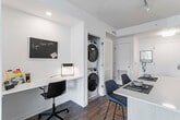 This single-floor residence showcases a contemporary open-concept design with warm hardwood flooring throughout. The kitchen is a standout feature, equipped with floor-to-ceiling light wood cabinetry, stainless steel appliances including a built-in microwave, dishwasher, gas range, and full-size refrigerator. A central island with a quartz countertop provides additional prep space and casual seating. A walk-in pantry offers generous storage directly off the kitchen. Adjacent to the kitchen, a dedicated workspace with a built-in desk and stacked in-unit washer and dryer adds everyday convenience.

The living area is bright and welcoming, with oversized windows fitted with roller shades that frame urban views and flood the space with natural light. Two bedrooms provide comfortable retreats; one features a large window with exterior views, while the primary bedroom offers ample room for furnishings. The bathroom is finished with dark hexagonal floor tiles, white subway tile surrounding the tub, a light wood vanity, and matte black fixtures, creating a clean, modern aesthetic throughout.