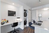 This single-floor residence showcases a contemporary open-concept design with warm hardwood flooring throughout. The kitchen is a standout feature, equipped with floor-to-ceiling light wood cabinetry, stainless steel appliances including a built-in microwave, dishwasher, gas range, and full-size refrigerator. A central island with a quartz countertop provides additional prep space and casual seating. A walk-in pantry offers generous storage directly off the kitchen. Adjacent to the kitchen, a dedicated workspace with a built-in desk and stacked in-unit washer and dryer adds everyday convenience.

The living area is bright and welcoming, with oversized windows fitted with roller shades that frame urban views and flood the space with natural light. Two bedrooms provide comfortable retreats; one features a large window with exterior views, while the primary bedroom offers ample room for furnishings. The bathroom is finished with dark hexagonal floor tiles, white subway tile surrounding the tub, a light wood vanity, and matte black fixtures, creating a clean, modern aesthetic throughout.