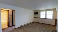 This single-floor space presents a clean and functional layout designed for practical everyday living. The main living area features wall-to-wall carpeting in a neutral tone, providing a comfortable underfoot feel throughout. White walls create a bright, open atmosphere, while dark wood baseboards add a subtle contrasting trim detail along the perimeter of the room. Climate control is well-addressed with a baseboard heating system running along the base of the exterior wall, complemented by a wall-mounted air conditioning unit, ensuring year-round comfort. A window allows natural light to filter into the space, offering a view of the surrounding greenery outside. A shelving ledge or half-wall detail near the window adds a modest architectural element to the room. An open doorway is visible, suggesting a connected layout that flows into an adjacent room or hallway, contributing to the overall sense of accessibility within the space. The overall ambiance is straightforward and move-in ready, suited for those seeking a no-frills, comfortable living environment.