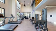 Located in Glendale, Arizona, this two-story fitness center offers a well-appointed workout facility designed to support a full range of exercise routines. The main floor commands immediate attention with its soaring double-height ceilings, oversized floor-to-ceiling windows, and an abundance of natural light that creates an open, energizing atmosphere. Light wood-tone flooring runs throughout, complementing the clean, contemporary palette of white and soft gray walls. A large ceiling fan anchors the central space, while wall-mounted TVs and full-length mirrors add both function and visual depth. This primary exercise room is equipped with a row of treadmills, stationary bikes, ellipticals, and a complete dumbbell rack, offering a thorough cardio and free-weight experience. A staircase with warm wood treads connects to the upper level, where a second exercise room provides a more intimate setting for specialized training. This upper-level room houses strength machines, a power rack, a leg press, and a Concept2 rowing ergometer, along with stability balls and additional free weights — ideal for resistance and cross-training workouts. A railed mezzanine overlooks the main floor below. Near the staircase on the ground level, a built-in cubby storage unit and dual water fountains set against a deep blue tile accent wall add practical amenities to the space.