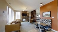 This thoughtfully designed single-floor communal space blends comfort and style into a welcoming gathering environment. The lounge is anchored by a striking stone fireplace wall, which serves as a natural focal point and adds rustic warmth to the room. A wall-mounted television above the fireplace makes the seating area ideal for both relaxed viewing and casual conversation. Plush armchairs and accent seating are arranged to encourage social interaction, complemented by soft carpeting underfoot.

A distinctive wooden lattice partition divides the space, offering a sense of separation between the lounge and dining areas while maintaining an open, airy feel. The dining zone features sturdy tables paired with industrial-style metal chairs, set against a bold terracotta-orange accent wall that energizes the room with color. Patterned bench seating and framed artwork add personality and visual interest.

Natural light filters in through slatted window treatments, while modern pendant and track lighting provide warm, layered illumination throughout. The combination of stone, wood, metal, and soft textiles creates a rich, multi-textured aesthetic. Overall, this space offers a versatile and inviting environment well-suited for socializing, dining, or simply unwinding in a beautifully curated setting.