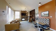 This thoughtfully designed single-floor communal space blends comfort and style into a welcoming gathering environment. The lounge is anchored by a striking stone fireplace wall, which serves as a natural focal point and adds rustic warmth to the room. A wall-mounted television above the fireplace makes the seating area ideal for both relaxed viewing and casual conversation. Plush armchairs and accent seating are arranged to encourage social interaction, complemented by soft carpeting underfoot.

A distinctive wooden lattice partition divides the space, offering a sense of separation between the lounge and dining areas while maintaining an open, airy feel. The dining zone features sturdy tables paired with industrial-style metal chairs, set against a bold terracotta-orange accent wall that energizes the room with color. Patterned bench seating and framed artwork add personality and visual interest.

Natural light filters in through slatted window treatments, while modern pendant and track lighting provide warm, layered illumination throughout. The combination of stone, wood, metal, and soft textiles creates a rich, multi-textured aesthetic. Overall, this space offers a versatile and inviting environment well-suited for socializing, dining, or simply unwinding in a beautifully curated setting.