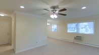This single-floor space presents a clean, functional living area designed with classic, understated details. The room is finished with crisp white crown molding that runs along the perimeter where the walls meet the ceiling, adding a refined architectural touch. A multi-blade ceiling fan with an integrated light fixture is centrally mounted, providing both air circulation and ambient lighting throughout the space.

The flooring is covered in neutral-toned carpet, contributing to a warm and quiet atmosphere underfoot. The walls are painted in a soft, light tone that enhances the sense of openness and pairs well with the white trim and baseboards running along the base of each wall.

Two windows allow natural light to enter the room, brightening the interior and offering views of the surrounding exterior greenery. A baseboard heating unit runs along the far wall, indicating a practical heating solution for the space. An additional window air conditioning unit is visible in one corner, providing climate control flexibility across seasons. Electrical outlets and a cable or data port are also present along the walls, supporting everyday functionality. Overall, the space offers a straightforward, well-maintained environment ready for use.