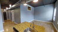 This single-floor space combines industrial design elements with modern finishes to create a stylish and functional environment. The kitchen area serves as a focal point, showcasing a striking granite island countertop with warm golden tones and a stainless steel sink with a pull-down faucet. The kitchen flows seamlessly into the open living area, creating a cohesive and spacious feel throughout.

The ceilings feature exposed silver ductwork and conduit, lending an industrial loft aesthetic to the space. Pendant lighting hangs from the dark ceiling, providing focused illumination and adding to the contemporary character. The light hardwood flooring runs throughout, offering a warm contrast to the cool gray-blue tones of the walls.

A small pass-through window is framed neatly into the wall, adding an architectural detail that enhances the open concept design. White baseboards provide a clean finishing touch throughout the space. The overall ambiance balances raw industrial character with refined modern touches, making it a versatile and visually engaging space suited for both everyday living and entertaining.
