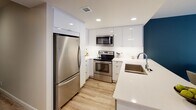 Located at 225 S Olive St in the heart of downtown Los Angeles, this thoughtfully designed single-floor unit offers a seamless blend of modern aesthetics and functional living. The open-concept main living area is anchored by light wood-style flooring and a bold teal accent wall, creating a striking visual focal point. The living space flows effortlessly into a dedicated dining area and a well-appointed kitchen featuring high-gloss white cabinetry, quartz countertops, stainless steel appliances including a refrigerator, range, and built-in microwave, and a peninsula with bar seating — ideal for casual dining or entertaining. The entryway makes a strong first impression with a decorative circular mirror wall piece and coordinating accent décor set against a deep teal wall. The bedroom is a serene retreat bathed in natural light, with direct access to a private balcony through floor-to-ceiling sliding glass doors, offering outdoor space with urban views. The bathroom is finished in clean white subway tile, complete with a glass-enclosed shower-tub combination and chrome fixtures. Recessed lighting throughout and neutral tones contribute to a bright, cohesive ambiance across the entire space.