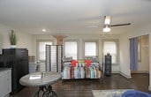 This single-floor unit offers an efficiently designed layout that seamlessly integrates living, sleeping, and dining functions within one open space. Dark hardwood flooring runs throughout, providing a warm, grounding contrast to the neutral gray and white walls.

The main living area is generously proportioned for its scale, featuring a ceiling fan with light fixture and multiple windows fitted with blinds that allow natural light to filter in. The space accommodates a sleeping area, a seating arrangement anchored by a patterned area rug, and a media console with storage cabinetry in a coordinating gray finish.

The kitchenette is positioned along one wall and includes gray lower and upper cabinetry, a double-basin sink, a black refrigerator, and a microwave. Dark hardwood flooring continues into this area, maintaining visual consistency. A small dining table with industrial-style seating completes the kitchen zone.

The bathroom is compact and functional, featuring a stand-up shower with a curtain enclosure, light tile flooring, and neutral wall finishes. Multiple interior doors provide access to a closet and the bathroom, contributing to a well-organized, practical floor plan overall.