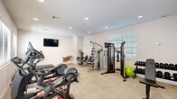 This fitness center in Lafayette, Louisiana, is a single-level space designed to support a comprehensive workout experience. The interior features clean white walls, recessed lighting, and light-toned flooring that combines carpet and hardwood-style sections, creating a bright and functional environment throughout.

The cardio area is well-stocked with multiple treadmills, elliptical trainers, and a Peloton stationary bike, all positioned to take advantage of natural light streaming in through several windows fitted with white horizontal blinds. A wall-mounted flat-screen television is centrally positioned, visible from most workout stations.

The strength training section includes a multi-station cable machine by Hoist, a leg press/extension unit, adjustable dumbbells on a rack, and a stability ball. A full-length wall mirror spans one side of the room, visually expanding the space and aiding in form correction during exercises.

Additional amenities include a built-in water fountain station along the back wall, providing convenient hydration access. A glass-paneled entry door allows visibility into the space. The overall design is clean, modern, and efficiently organized to maximize usability within the available footprint, making it a practical and well-appointed fitness facility.