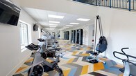 This two-story fitness center offers a well-equipped and visually dynamic environment designed to support a full range of workout routines. The main floor is expansive, featuring an impressive lineup of cardio equipment including spinning bikes, ellipticals, treadmills, and a rowing machine, as well as dedicated strength training stations and cable machines along the far wall. The space is anchored by a striking chevron-patterned floor in bold shades of gold, blue, gray, and natural wood tones, adding an energetic visual dimension to the workout environment. Bright recessed panel lighting in the drop ceiling ensures the space is evenly and thoroughly lit throughout. Motivational wellness-themed typography adorns the accent wall, reinforcing the health-focused atmosphere. Wall-mounted screens are positioned for convenient viewing during cardio sessions. A mezzanine level is visible overhead, accessible via a staircase, providing additional space for fitness activities. The overall ambiance is modern, clean, and inviting, with an open layout that allows for comfortable movement between equipment zones and accommodates a variety of fitness preferences and training styles.